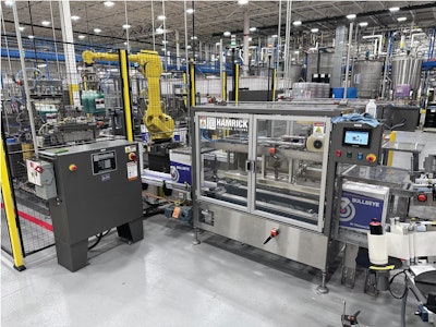 Un brazo robótico de cinco ejes toma dos botellas llenas a la vez y las ubica en las cajas corrugadas con particiones. Las cajas se cierran, encintan, y reciben la etiqueta antes de llegar al paletizador.