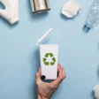 Hand holding white recycling container surrounded by plastic bottles and packaging waste