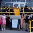 Group of officials and dignitaries at a ceremonial plaque unveiling event with team members in yellow and black uniforms observing from above.
