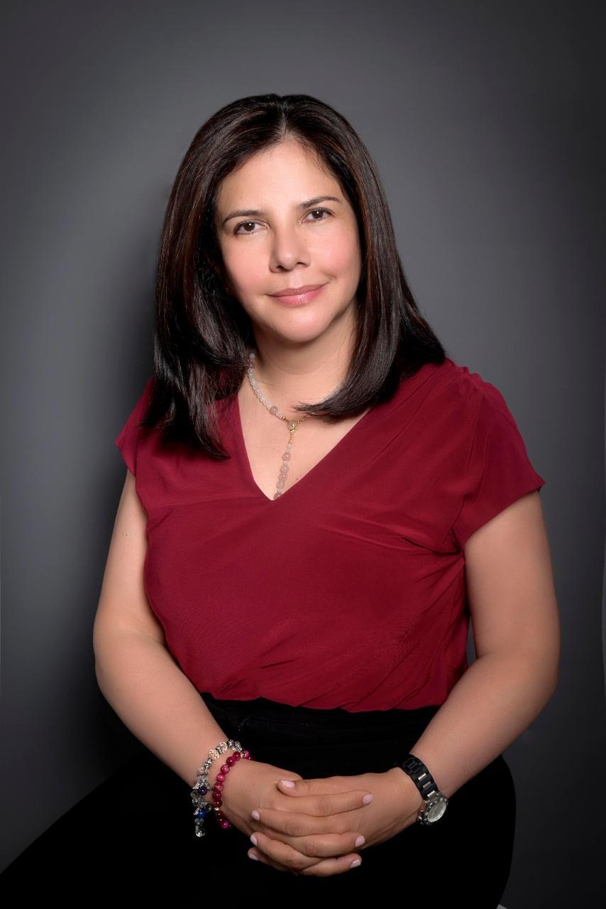 Professional headshot of woman wearing burgundy top and black skirt with jewelry against gray background