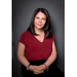 Professional headshot of woman wearing burgundy top and black skirt with jewelry against gray background