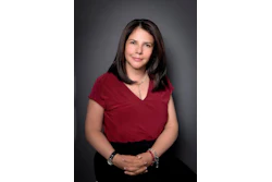 Professional headshot of woman wearing burgundy top and black skirt with jewelry against gray background