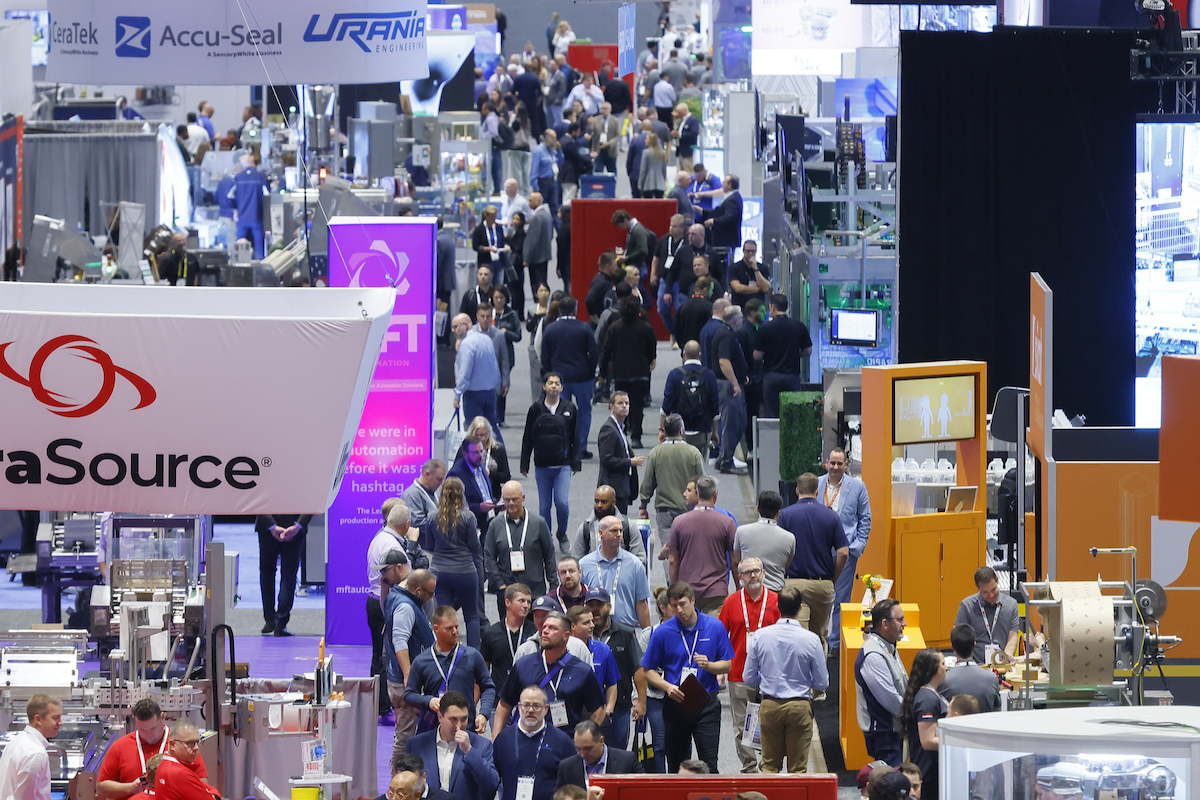 Crowded trade show exhibition floor with multiple company booths, signage, and attendees networking