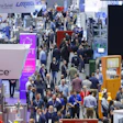 Crowded trade show exhibition floor with multiple company booths, signage, and attendees networking
