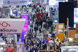 Crowded trade show exhibition floor with multiple company booths, signage, and attendees networking