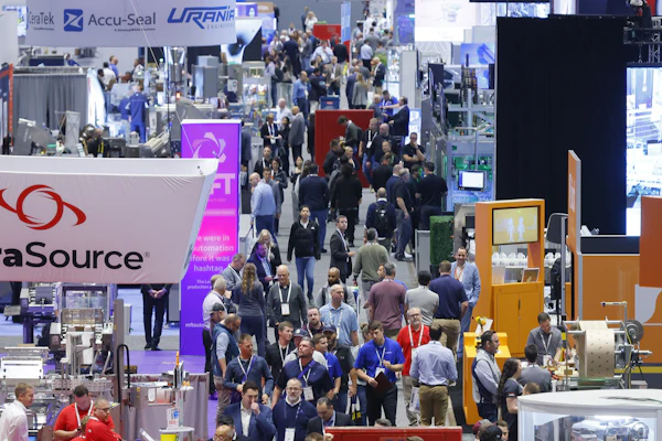 Crowded trade show exhibition floor with multiple company booths, signage, and attendees networking