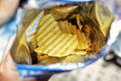 Open bag of ridged potato chips with golden-yellow wavy crisps visible inside blue and white packaging